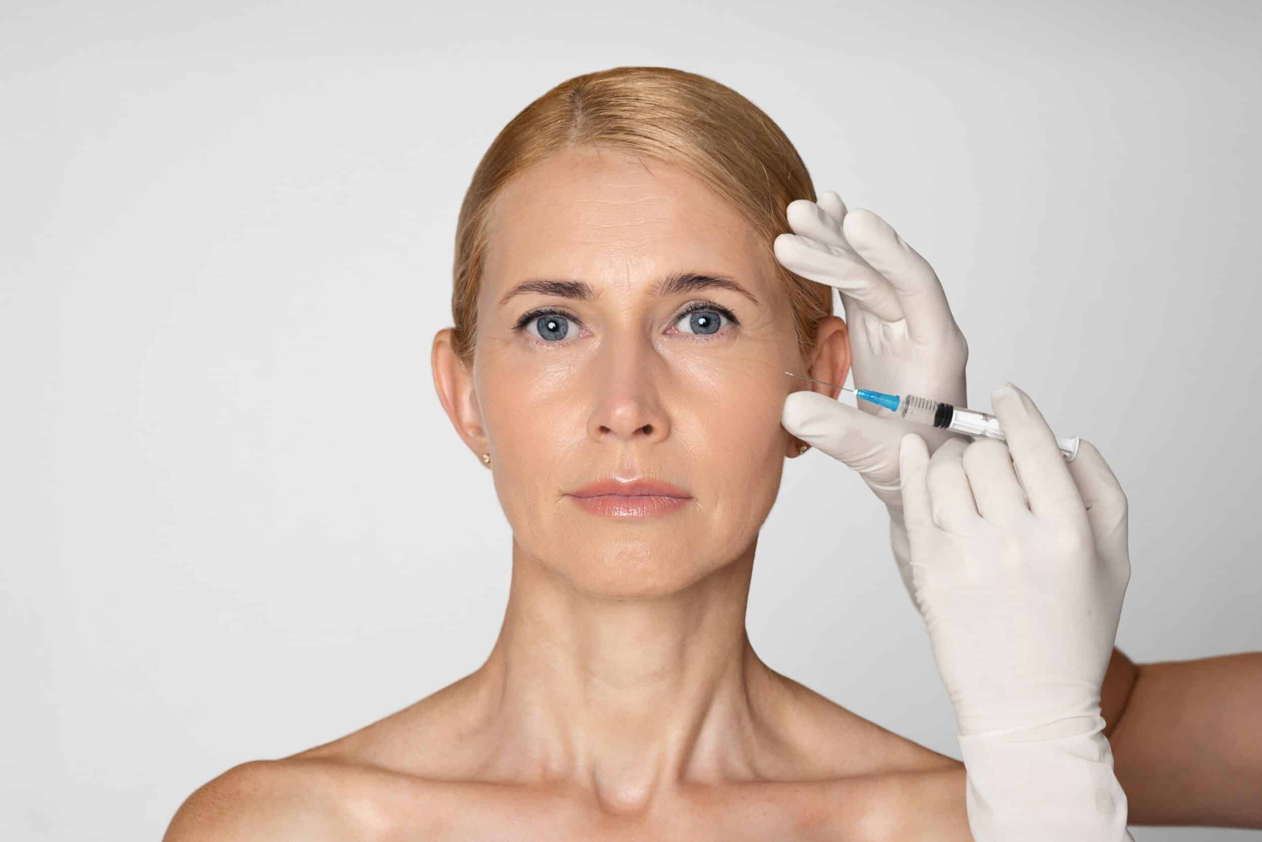 A doctor's gloved hand holds a syringe to administer dermal fillers in a woman's cheeks